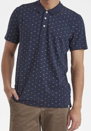 Mężczyzna w granatowej krótkiej koszulce polo z białymi abstrakcyjnymi wzorami i khaki spodniach, stojący z jedną ręką w kieszeni.