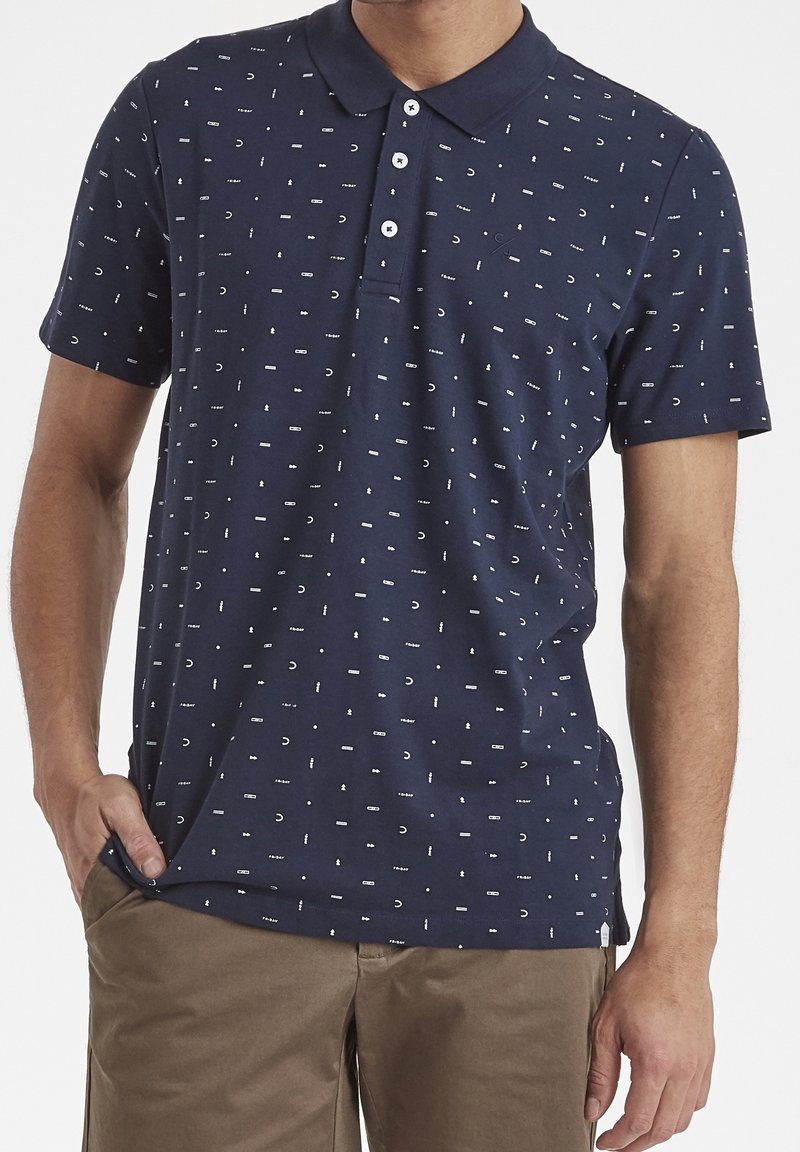 Homme portant un polo à manches courtes bleu marine avec des motifs abstraits blancs et un pantalon kaki, debout avec une main dans la poche.