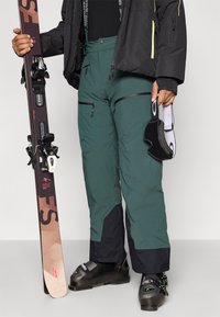 Calças de esqui em verde escuro com acabamentos em preto, com bolsos com fecho, corte ajustado e material à prova d'água, usadas com equipamento de esqui.