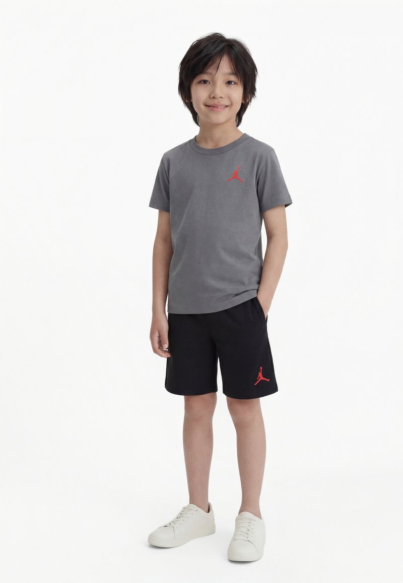 Νέος αγόρι όρθιος, φορά γκρι μπλούζα και μαύρο σορτς με κόκκινα λογότυπα Jordan, και λευκά αθλητικά παπούτσια σε απλό λευκό φόντο.