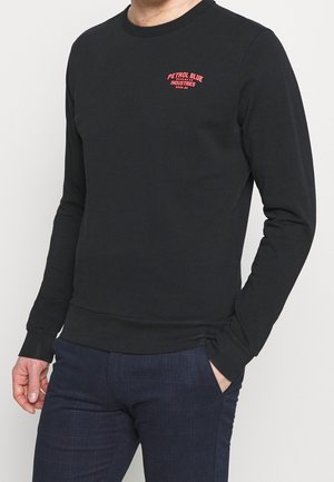 Mann trägt schwarzes Langarmshirt mit rotem "Petrol Blue Industries"-Logo auf der Brust und dunkelblaue Hose, eine Hand in der Tasche.