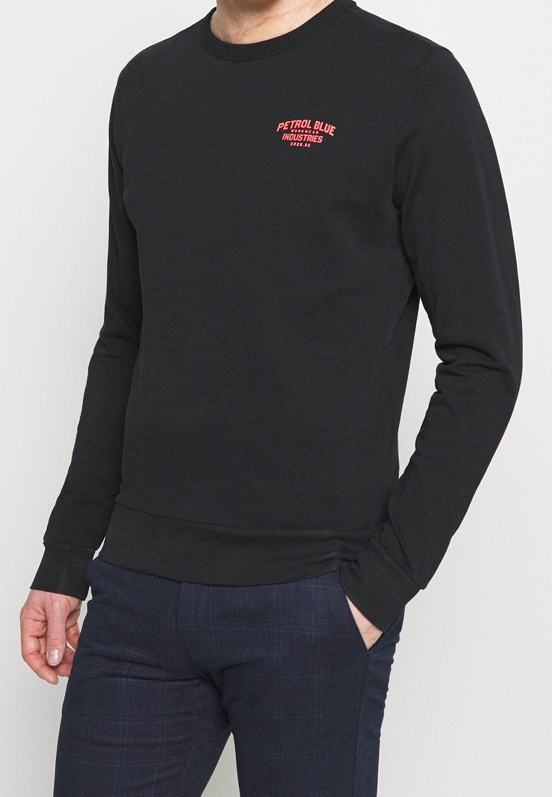 Homme portant une chemise noire à manches longues avec le logo rouge « Petrol Blue Industries » sur la poitrine et un pantalon bleu foncé, une main dans la poche.