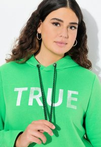 Felpa verde brillante con lettering bianco "TRUE". Presenta coulisse con dettagli neri e una tasca a marsupio. Tessuto morbido con vestibilità rilassata.