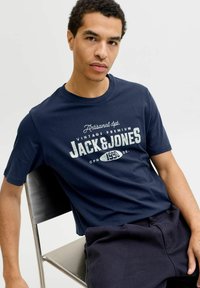Camiseta de algodón azul marino con mangas cortas, que presenta un logo impreso en blanco de estilo vintage. El modelo lleva pantalones cortos oscuros y está sentado en una silla.