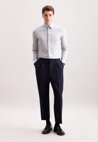 Uomo in piedi con le mani in tasca, indossa una camicia da uomo grigia chiara, pantaloni in navy con pieghe, calze nere e scarpe eleganti nere.