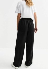 Schwarze weite Hosen mit elastischem Bund, aus glattem Stoff, kombiniert mit einem weißen T-Shirt und weißen Sneakers.