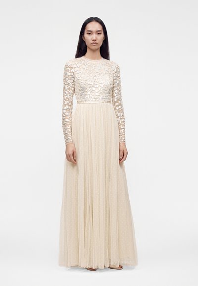 Femme portant une longue robe crème avec un corsage transparent brodé de fleurs et des manches longues, debout devant un fond clair uni.