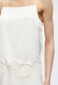 Haut blanc sans manches avec fines bretelles et bordure en dentelle florale le long du bord inférieur, porté sur la partie supérieure du corps d'une personne.