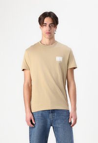 Beige cotton t-shirt with short sleeves and a round neckline, featuring a small printed label on the left chest. Worn with blue jeans.