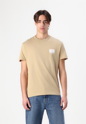 Beige cotton t-shirt with short sleeves and a round neckline, featuring a small printed label on the left chest. Worn with blue jeans.