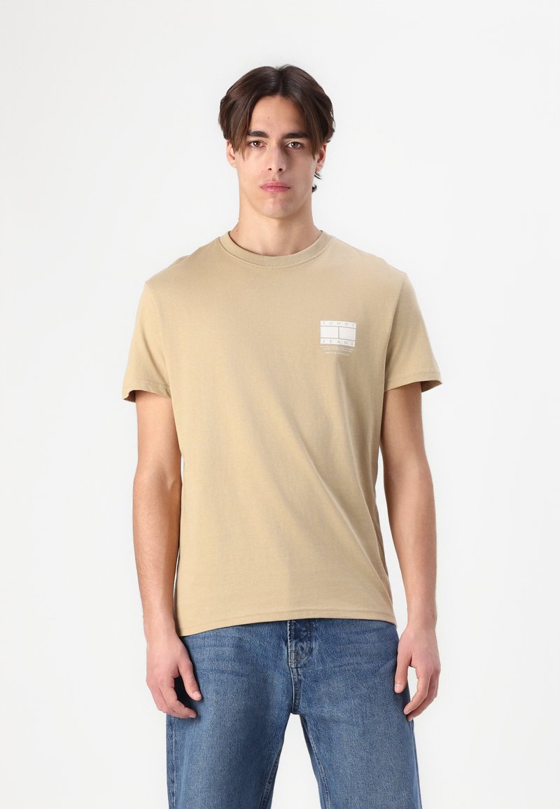 Beige cotton t-shirt with short sleeves and a round neckline, featuring a small printed label on the left chest. Worn with blue jeans.