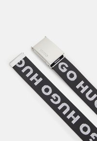 Cintura nera in tela con bold lettering bianco "HUGO", dotata di una fibbia rettangolare in metallo argentato e estremità piatte.