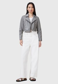Giubbotto di pelle grigio corto con tasche con zip, abbinato a pantaloni bianchi a vita alta e sandali argentati. Texture liscia, design minimalista.