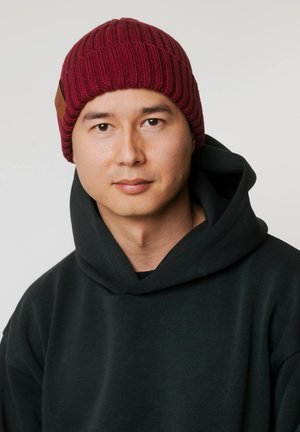 Bordowa dzianinowa czapka z prążkowaną fakturą i brązową skórzaną naszywką, zestawiona z ciemną bluzą z kapturem. Zbliżenie na głowę i ramiona.