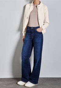 Chaqueta beige claro con cremallera sobre una camisa marrón claro, combinada con vaqueros anchos de mezclilla azul oscuro y zapatillas blancas.