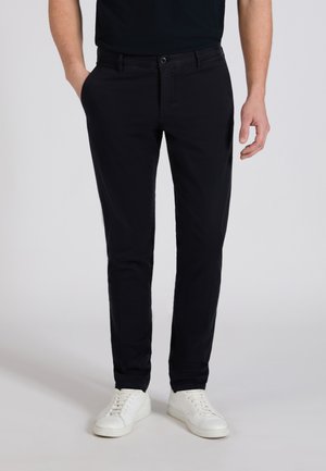 Man draagt smalle zwarte broek met handen in de zakken en witte sneakers tegen een effen lichte achtergrond.