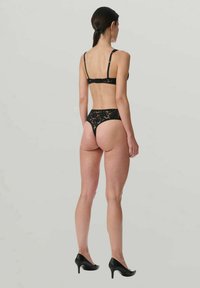 Femme se tenant de dos, portant un soutien-gorge en dentelle noire et un string assorti, avec des escarpins noirs sur un fond uni.