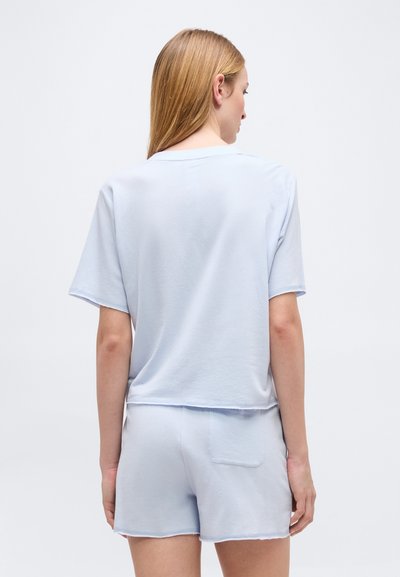 Γυναίκα με μακριά μαλλιά που φοράει ανοιχτό μπλε τοπ με κοντά μανίκια και ταιριαστό σορτς, στεκόμενη με την πλάτη προς την κάμερα.