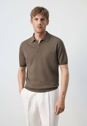 Hombre con cabello ondulado corto, vestido con una camisa de color verde oliva de manga corta y cuello, y pantalones blancos, de pie frente a un fondo claro y sencillo.