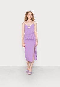 Vestido bodycon lavanda com alças finas, recorte frontal, detalhe franzido na lateral e fenda alta na coxa; combinado com sandálias de salto tecidas a condizer.