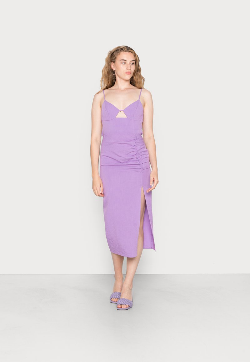 Vestido bodycon lavanda com alças finas, recorte frontal, detalhe franzido na lateral e fenda alta na coxa; combinado com sandálias de salto tecidas a condizer.