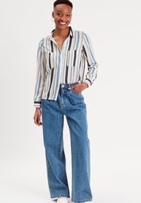 Chemise à rayures verticales en crème, bleu et noir ; deux poches à l'avant. Associée à un jean en denim bleu ample et des chaussures blanches.