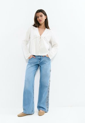 Femme aux longs cheveux bruns porte un chemisier blanc à manches longues boutonné et un jean bleu clair à jambes larges avec une broderie circulaire sur les côtés.