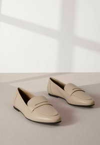 Mocassini in pelle beige con punta arrotondata e una cintura decorativa sulla parte anteriore. Suole nere piatte e superficie liscia.