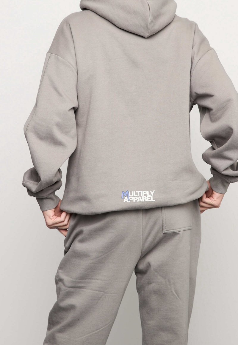 Persona que lleva una sudadera con capucha gris claro y pantalones a juego, manos sosteniendo el dobladillo de la sudadera, logotipo de "Multiply Apparel" visible en la parte baja de la espalda.