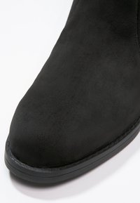 Bottine en daim noir avec bout arrondi et détail de semelle cousue sur fond blanc.