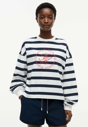 Persoon draagt een wit met marineblauw gestreepte trui met rode tekst en marineblauwe korte broek, staand met één hand in de zak tegen een witte achtergrond.