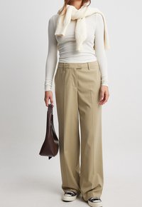 Kvinna iklädd beige vida byxor, vit långärmad topp med en krämfärgad tröja över axlarna, håller en mörkbrun handväska, bär sneakers.