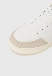 Zapatilla blanca con acentos de ante gris claro. Presenta puntera perforada, cordones redondos y una suela de goma texturizada. Diseño suave y limpio.