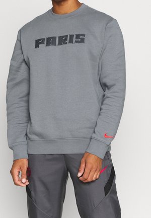 Mężczyzna w szarej bluzie z napisem "PARIS" i szarych spodniach z czerwonymi logo Nike na rękawie i nogawce.