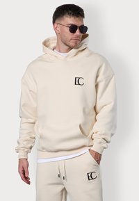 Beige Kapuzenpullover mit einer vorderen Tasche und gesticktem Logo auf der Brust. Weicher Stoff, lockere Passform, kombiniert mit passenden Jogginghosen.