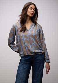 Femme aux longs cheveux bruns portant un chemisier ample à motifs bleus et bruns et un jean bleu foncé, debout devant un fond gris uni.