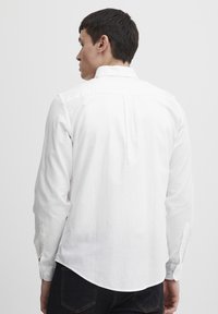Camisa blanca de manga larga con cuello abotonado, dobladillo curvado y un pliegue en la parte trasera. Tejido texturizado con puños de botón y aberturas laterales.