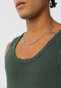 Kaki groene ribbel tanktop met een ronde hals en rauwe randen, voorzien van dunne schouderbanden en een gestructureerde stof. Zilveren ketting met een hanger zichtbaar.