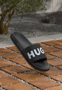 Sorte slidesandaler med en tekstureret fodseng og en bred rem med "HUGO" i fede hvide bogstaver, sat mod en mønstret fliseoverflade.
