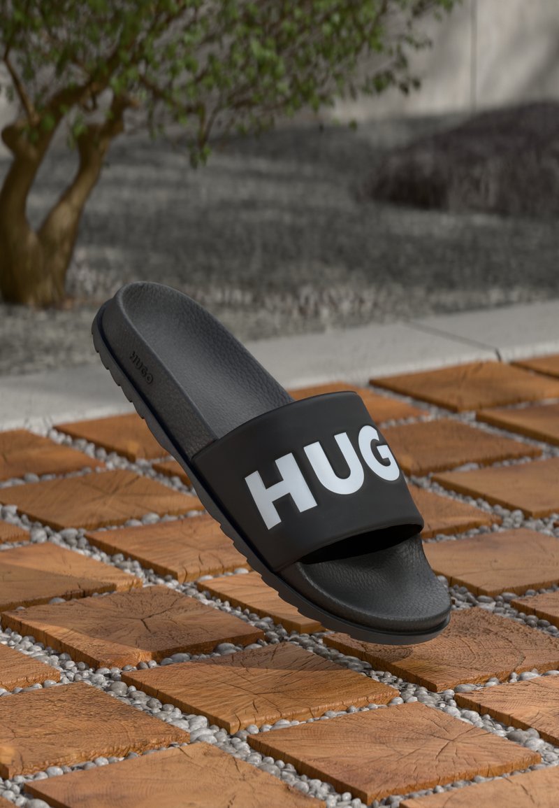 Sorte slidesandaler med en tekstureret fodseng og en bred rem med "HUGO" i fede hvide bogstaver, sat mod en mønstret fliseoverflade.