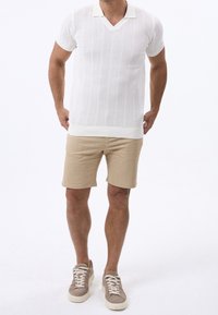Mand iført en hvid kortærmet strik polo, beige shorts og beige sneakers står imod en hvid baggrund.