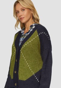 Strickjacke mit einem grün-blauen geometrischen Muster, strukturiertem Stoff, V-Ausschnitt und großen Knöpfen. Über einem karierten Hemd getragen.