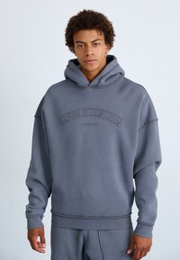Sweatshirt à capuche gris avec une coupe décontractée. Présente le texte "Alpha Industries" brodé en arc sur la poitrine. Le tissu a une texture douce.