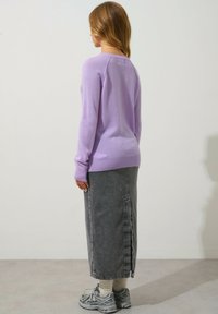 Maglione lilla con maniche lunghe, abbinato a una gonna di jeans grigia con uno spacco posteriore. Scarpe sportive grigie con dettagli testurizzati completano il look.
