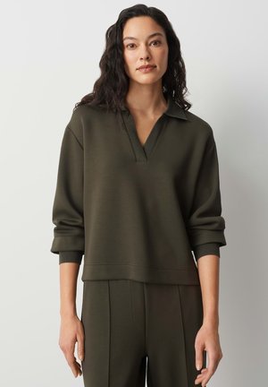 Femme aux longs cheveux foncés portant un pull à col en vert olive et un pantalon assorti sur un fond clair uni.