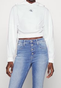 Biała krótka bluza z kapturem ze sznurkiem i haftowanym logo. Do tego dżinsy z wysokim stanem w kolorze niebieskim, z pięcioma przednimi guzikami i kieszeniami.