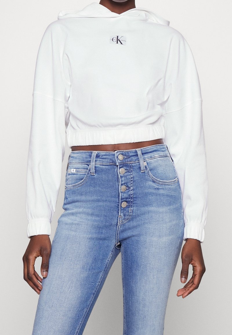 Biała krótka bluza z kapturem ze sznurkiem i haftowanym logo. Do tego dżinsy z wysokim stanem w kolorze niebieskim, z pięcioma przednimi guzikami i kieszeniami.