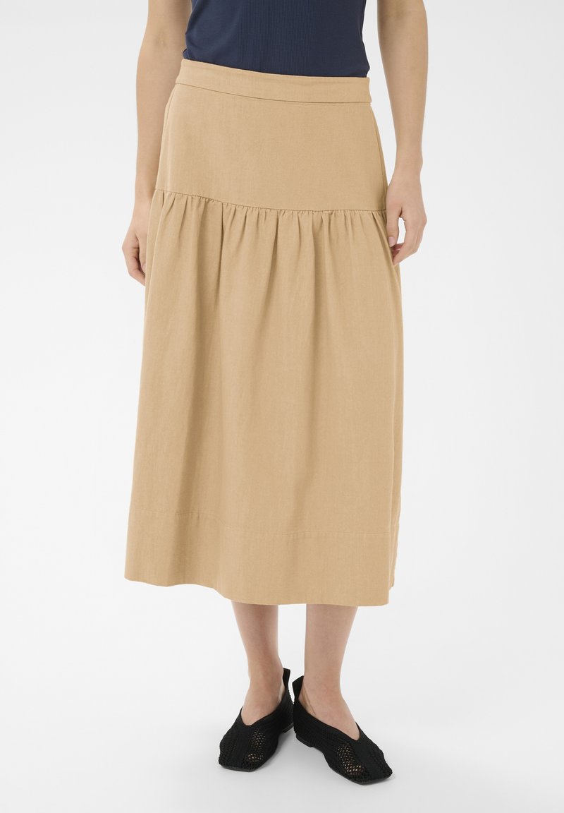 Femme portant une jupe midi beige avec une couture froncée et des chaussures plates noires en tissu tissé, debout devant un fond blanc.