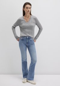 Grau gestrickter Pullover mit V-Ausschnitt, kombiniert mit hellblauen Schlagjeans und hellen Sneakers. Glatter Stoff mit subtiler Textur.