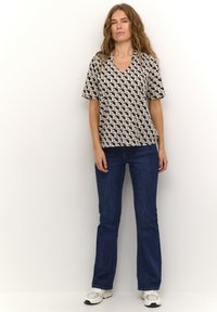 Top a maniche corte e scollatura a V in un tessuto beige con un motivo geometrico a cubi. Abbinato a jeans svasati blu scuro e sneakers bianche.